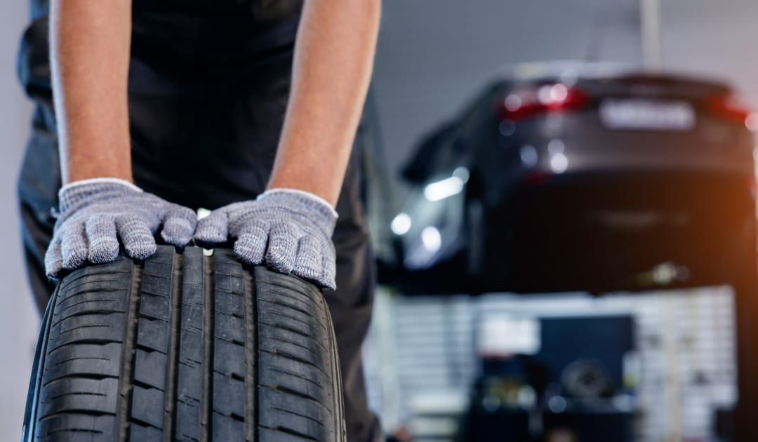 Close-up of a mechanic wearing grey work gloves inspecting the tread depth and wear pattern of an unmounted tire in an auto shop.